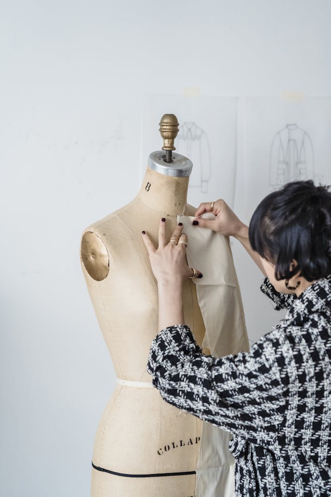 Side view of crop unrecognizable female in trendy outfit trying on white fabric on mannequin in light workshop